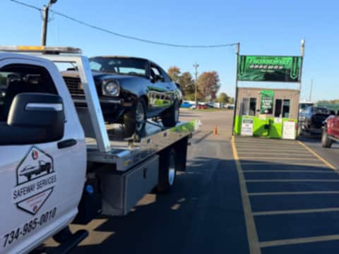 Black classic car strapped down on a Safeway Services flatbed, arriving at a drag strip entrance in Milan, Michigan