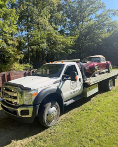 Maroon Shelby Cobra replica loaded on a Safeway Services flatbed tow truck along a tree-lined gravel drive