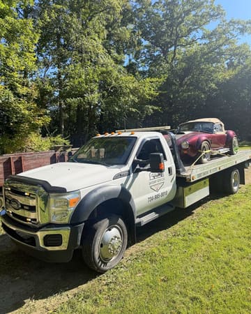 Maroon Shelby Cobra replica loaded on a Safeway Services flatbed tow truck along a tree-lined gravel drive